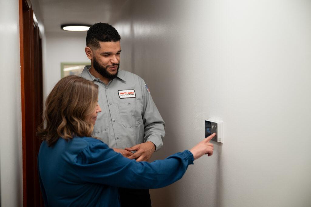 Technician showing a homeowner how to adjust a smart thermostat on a white wall in a hallway.